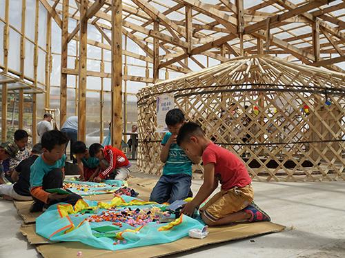 A group of children sit on the floor of a wood-framed and glass-clad structure. The children are interacting with colorful materials.